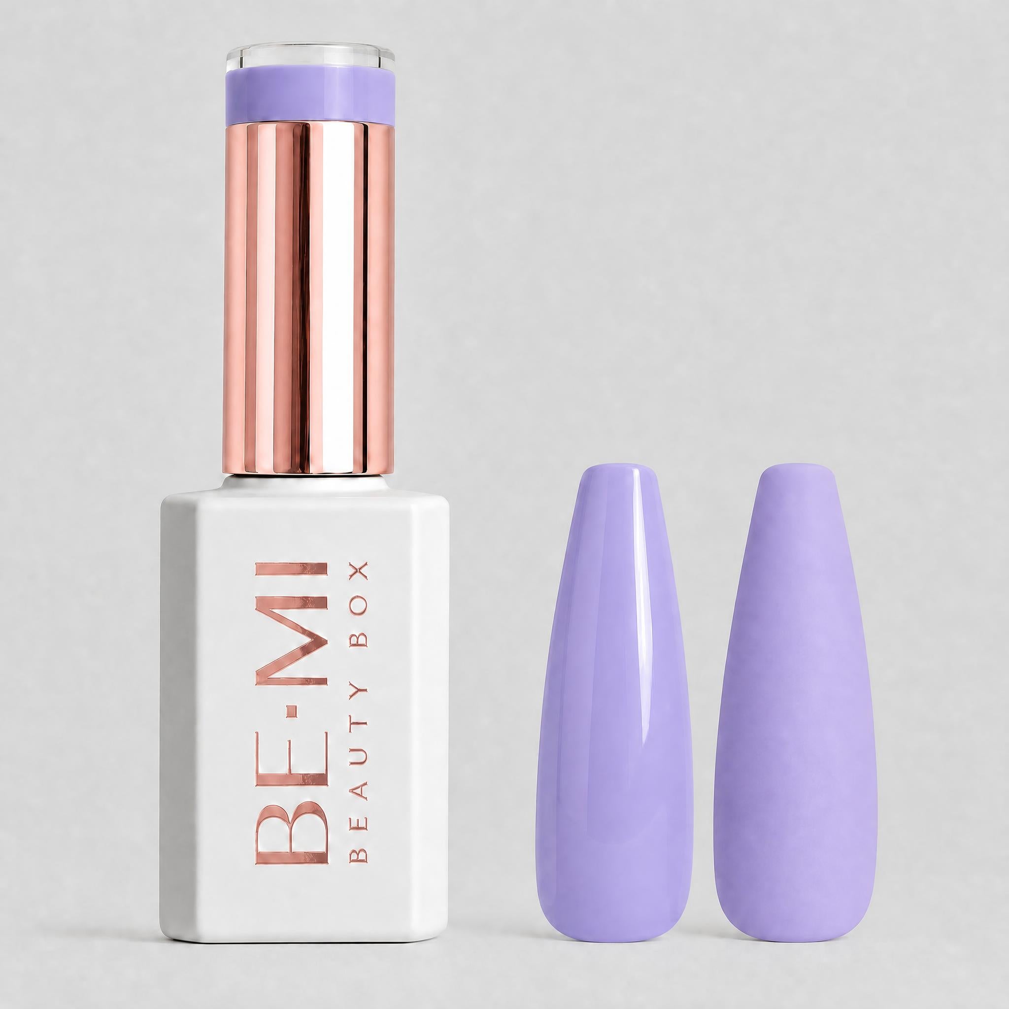 A bottle of BE-MI Beauty Box nail polish with a metallic rose gold cap and a white base, shown next to two artificial nail tips painted in a light lavender purple shade, on a neutral gray background.