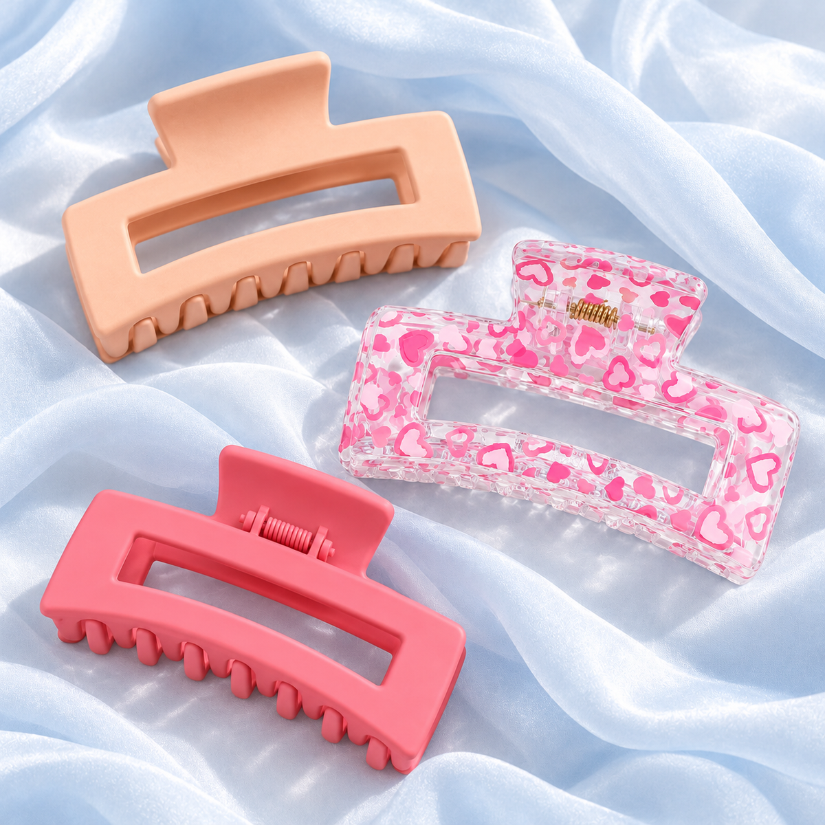 Coconoir Beauty large hair claw clips in Pink Hearts, Peach, and Coral resting on softly folded pale-blue fabric.