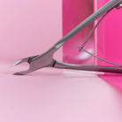 A close-up of a metallic nail clipper or cuticle tool resting diagonally on a pink surface with a matching pink background.