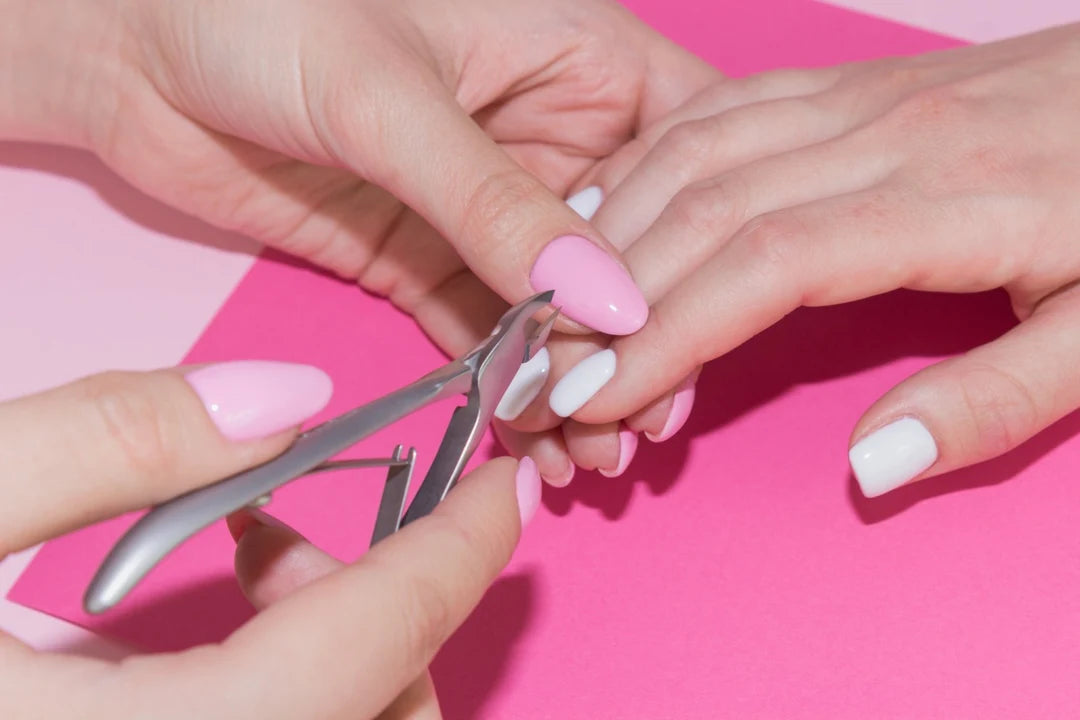A close-up of manicure tools being used on a person's fingernails, with one nail clipped while another is painted pink and others are white, against a bright pink background.